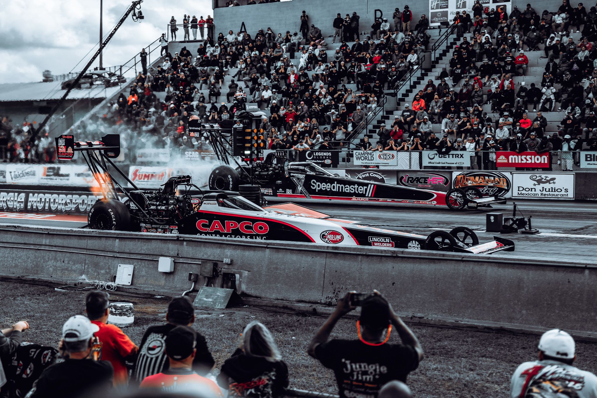 Two NHRA Top Fuel dragsters launching side by side through smoke and flame
